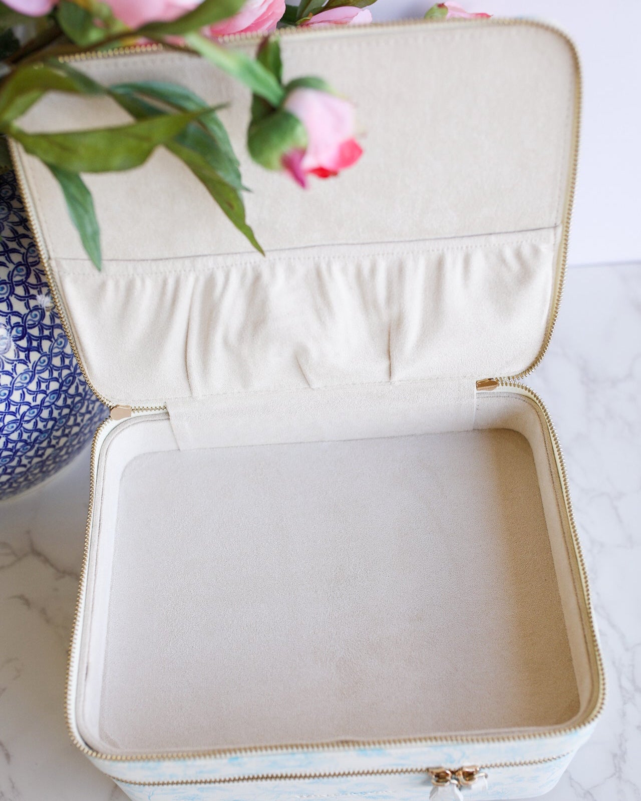 Beige jewelry box with gold zipper on a marble surface with pink flowers.